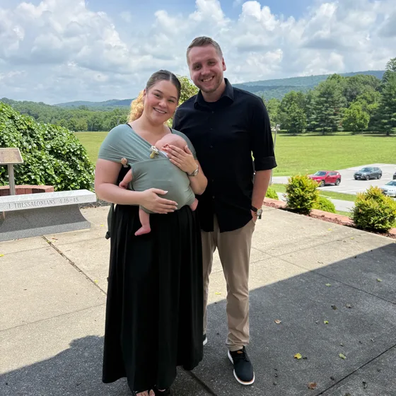 Tim and Elise Morgan with their baby in a carrier, standing outdoors on a sunny day with green hills behind them